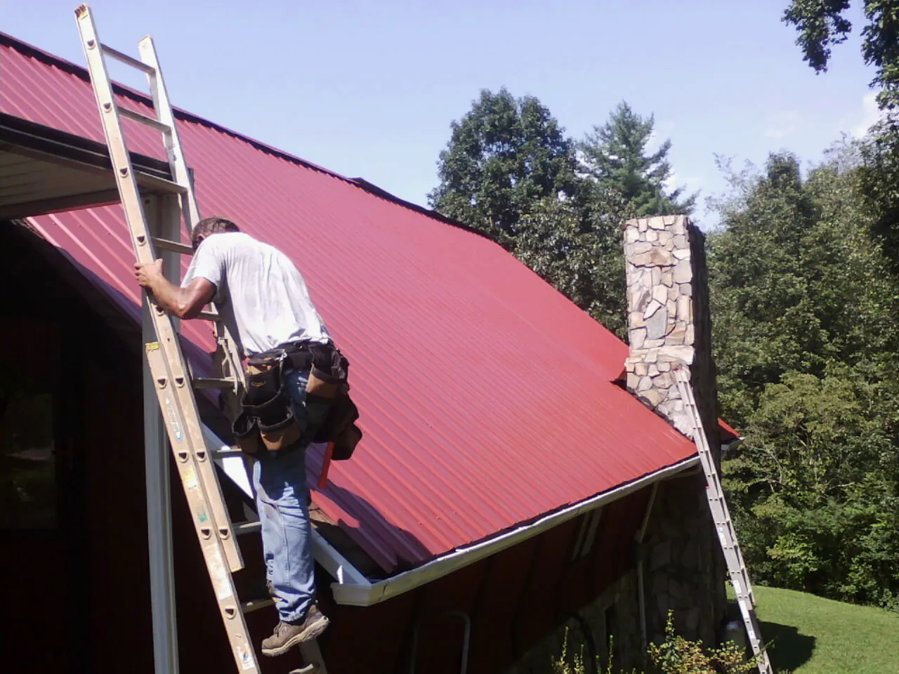 Roofer on a ladder beside a red metal roof and stone chimney — Soffit & Fascia Repair by Roofing Brothers in Duff